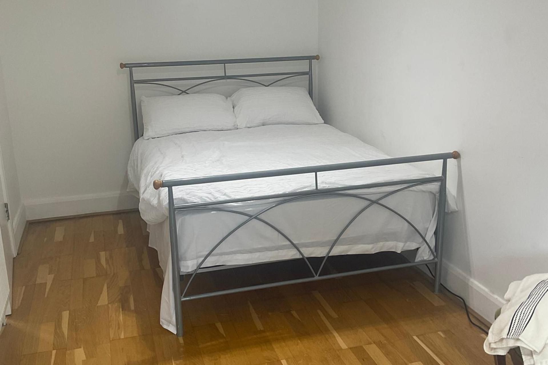 Een bed met metalen frame en witte beddengoed in een kamer met houten vloer en witte wanden.