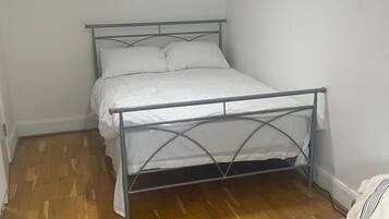 Een bed met metalen frame en witte beddengoed in een kamer met houten vloer en witte wanden.
