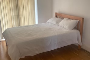 Habitación individual con una cama de una sola plaza, cabecera de madera, ropa de cama blanca y una ventana con persianas.
