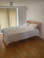 Un dormitorio con estructura de cama de madera, ropa de cama blanca y una ventana con persianas.