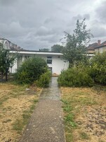 Un sendero que lleva a un edificio blanco con una ventana y una puerta, rodeado de vegetación y casas residenciales al fondo.