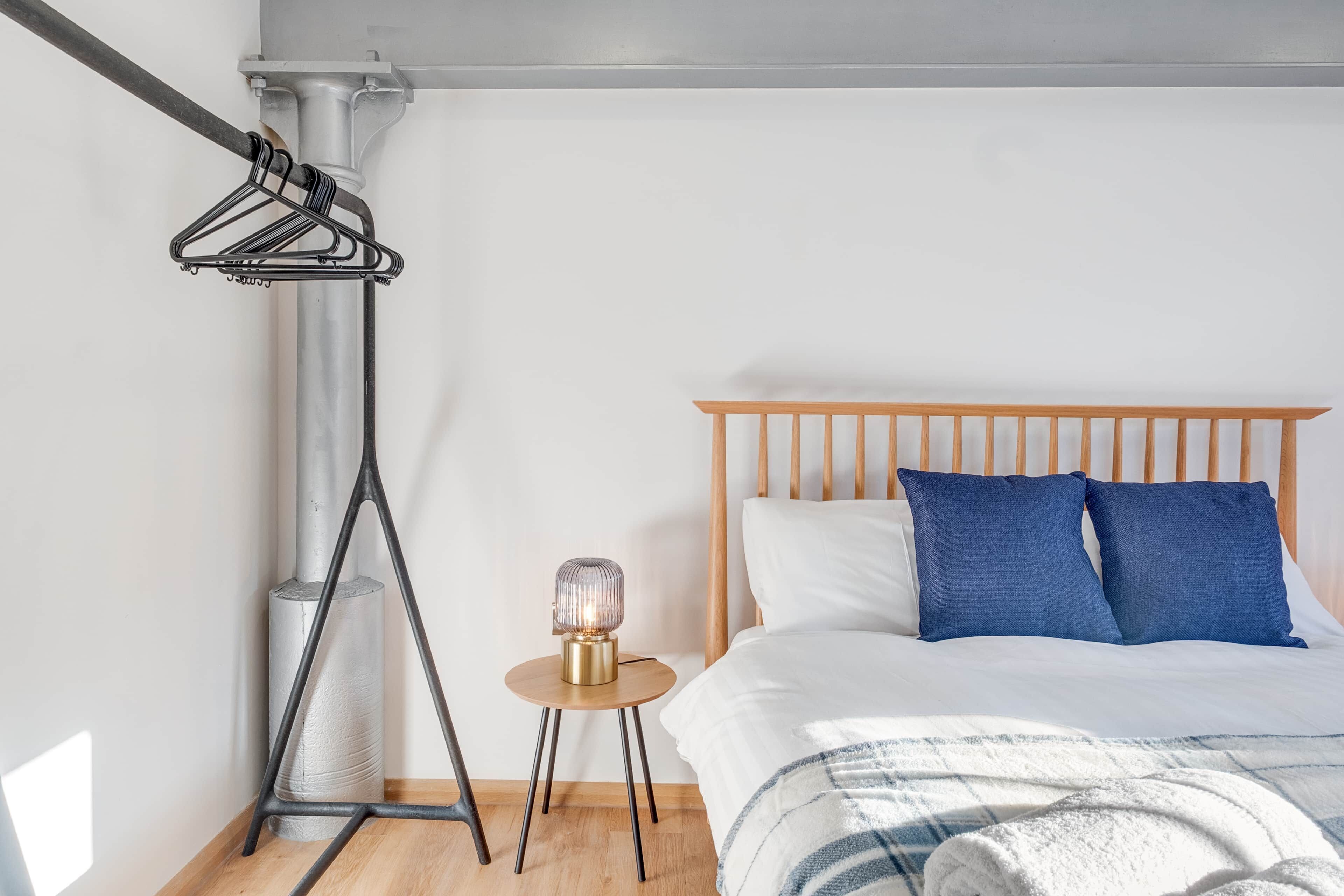 A bedroom with a wooden bed, white bedding, a small table with a lamp, and a clothing rack.