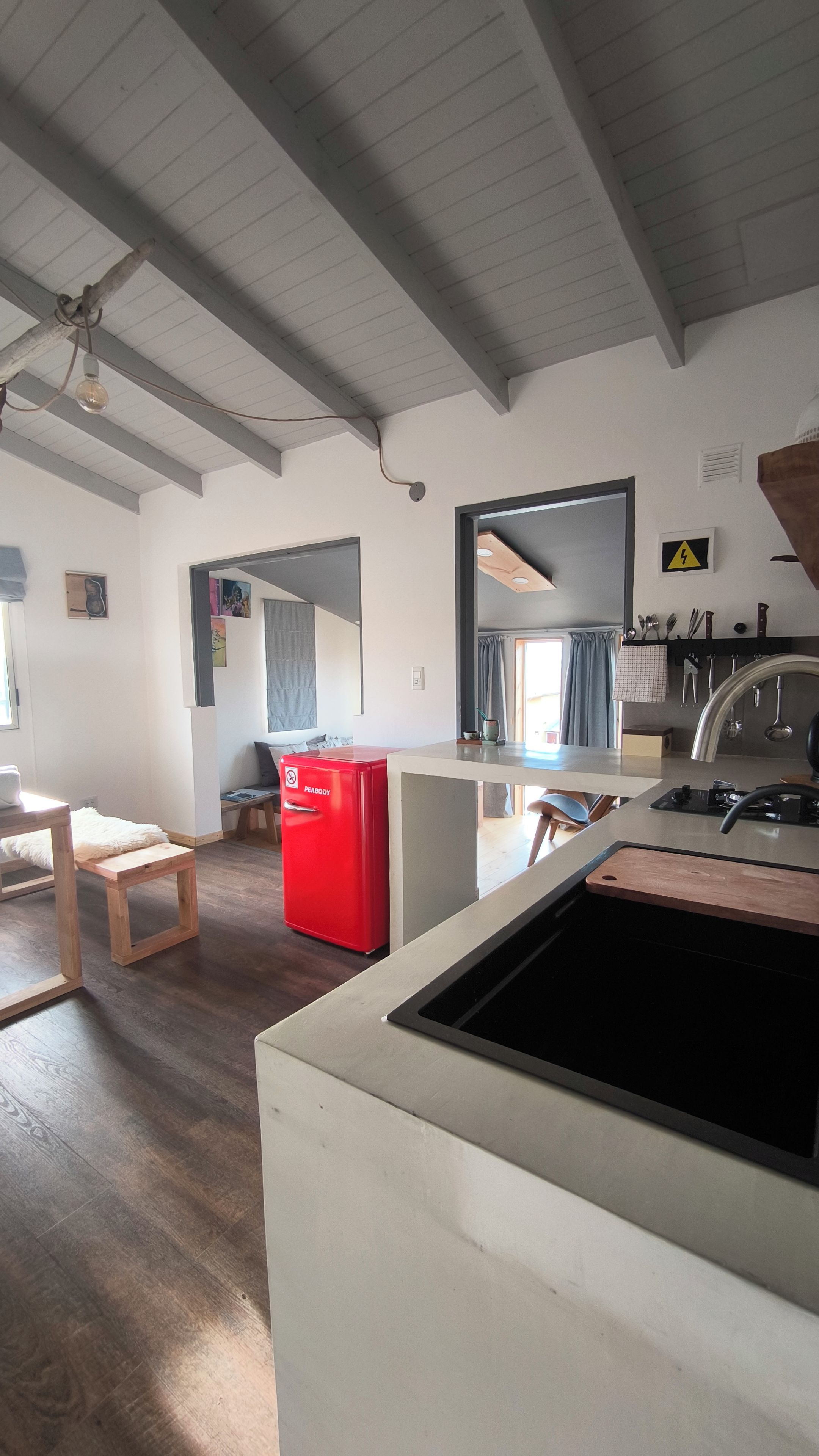 Cozinha com pia, eletrodoméstico vermelho e espelho. Há uma janela com cortinas e um banco de madeira.