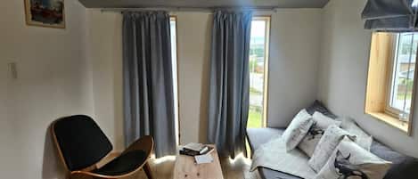 A room with a wooden table, a black chair, a sofa with patterned cushions, and a window with curtains.