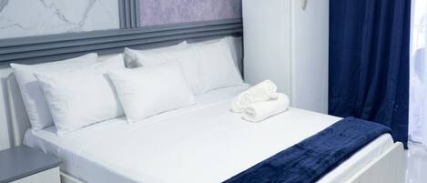 A hotel room with a bed, white pillows, a blue blanket, a white wardrobe, and a nightstand.