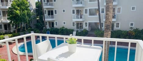 Un balcón de hotel con piscina, mobiliario blanco y vistas al exterior del edificio.
