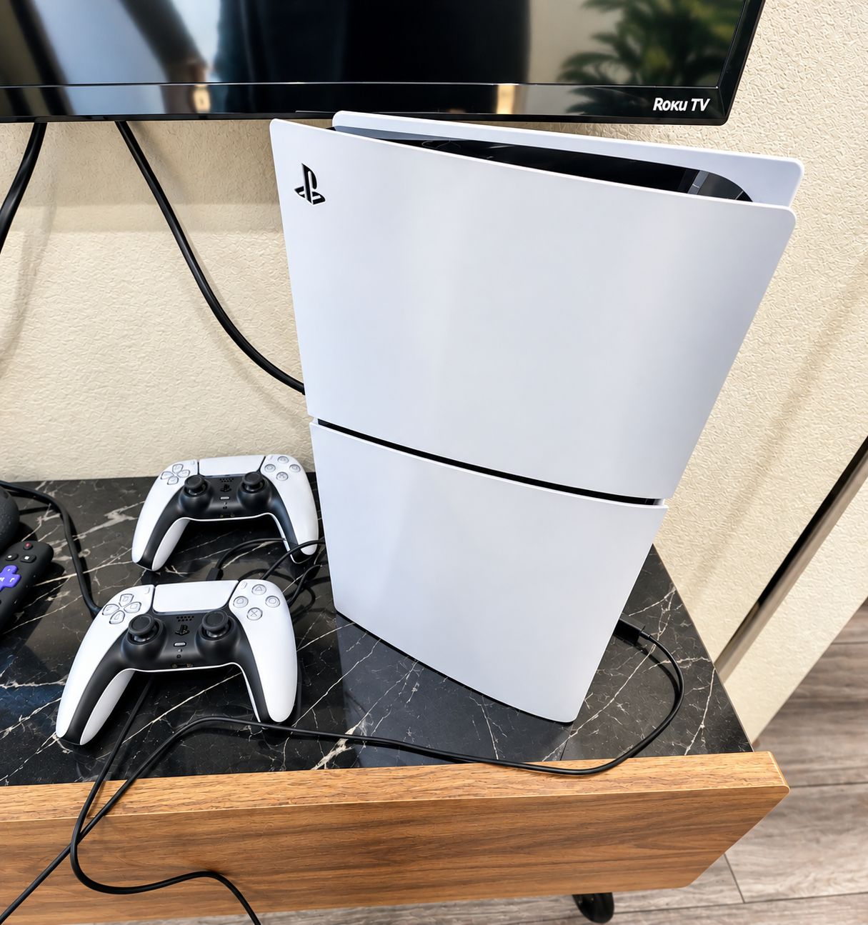 A white PlayStation 5 console on a marble table with a wooden side table, a black TV, and a PlayStation controller.
