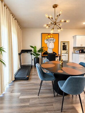 A modern dining area with a round table, chairs, a treadmill, and a large painting on the wall.