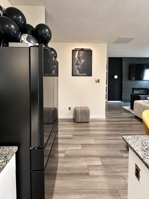 A modern kitchen with a black refrigerator, wooden flooring, a grey ottoman, and a dining area with a granite countertop. - Luxury 5BR Furnished Spacious Dallas Retreat. Short & Extended Stays. Sleeps 16! (Glenn Heights)