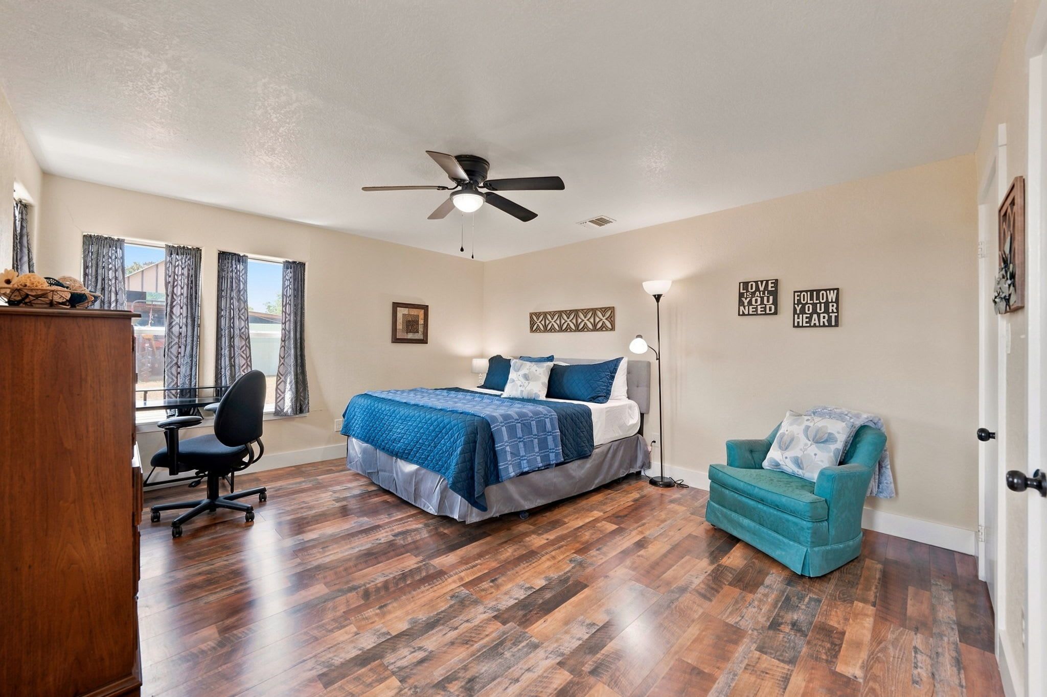 A bedroom with a bed, a desk, a chair, a ceiling fan, and a window with curtains.