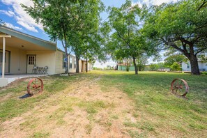A grassy yard with two large trees, a building with a porch, and a fence in the background.