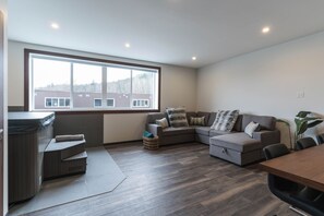 A modern living room with a sectional sofa, a small table, and a large window offering a view of buildings. - The Timber Den (Revelstoke)