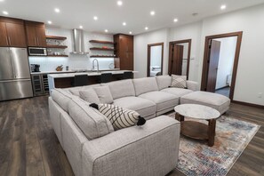 A modern living room with a sectional sofa, a round coffee table, and a kitchen area with stainless steel appliances. - The Timber Den (Revelstoke)