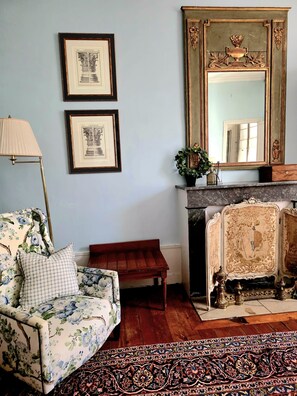 Una habitación con un sofá estampado con motivos florales, un pequeño banco de madera, una chimenea con detalles ornamentales y dos dibujos arquitectónicos enmarcados en la pared.