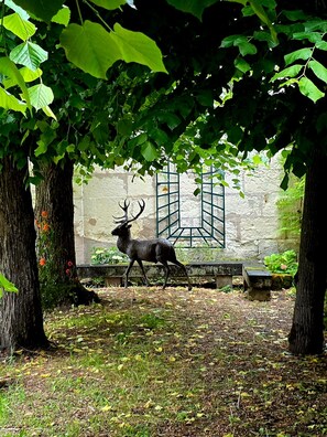 Eine Hirschstatue in einer schattigen Umgebung mit Bäumen und einer Steinmauer im Hintergrund.