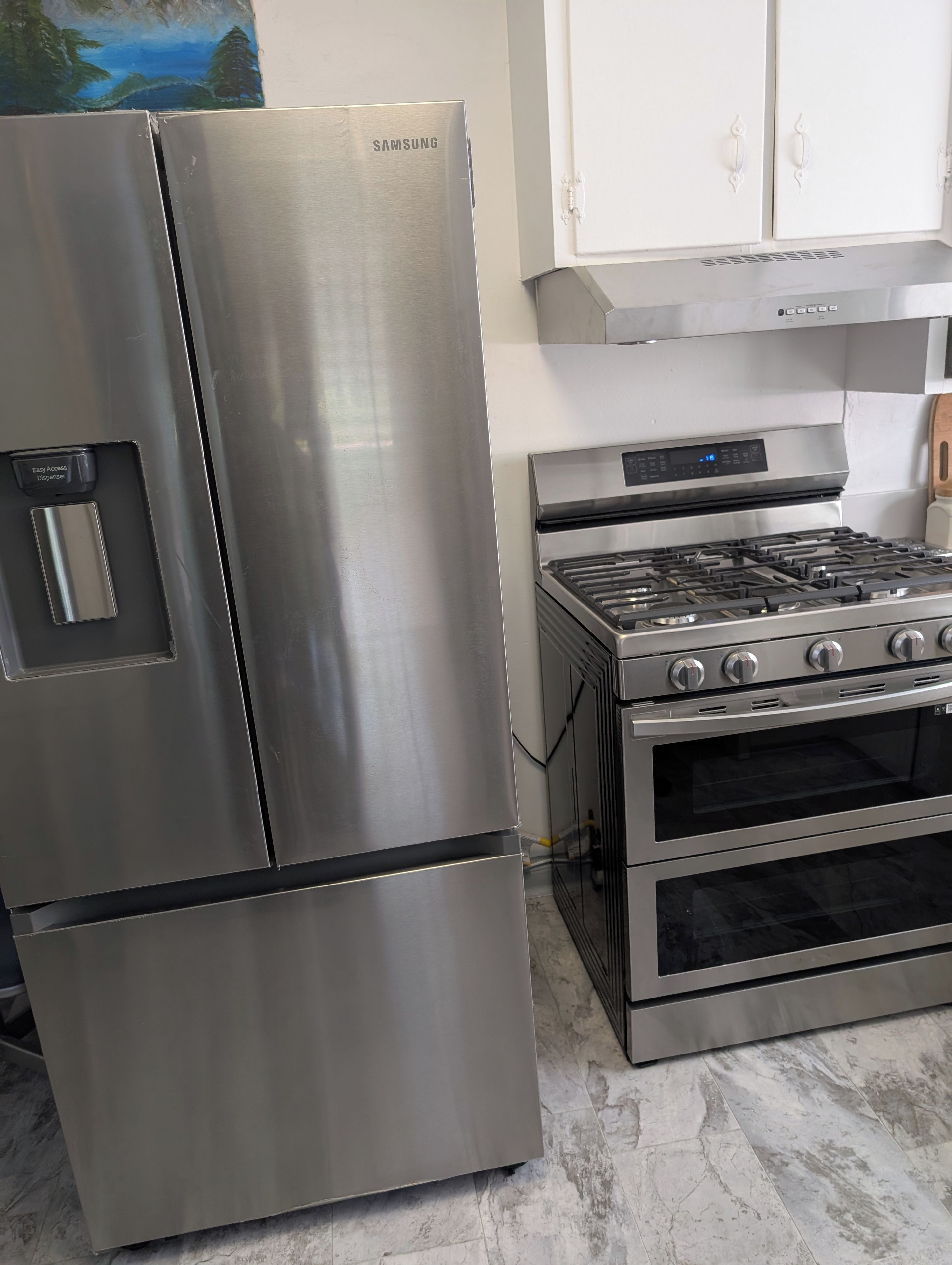 A stainless steel Samsung refrigerator and a built-in gas range with oven, in a kitchen with white cabinets and marble flooring.