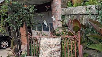 A wrought iron bench with a mosaic cushion in a garden area with potted plants and a building in the background.