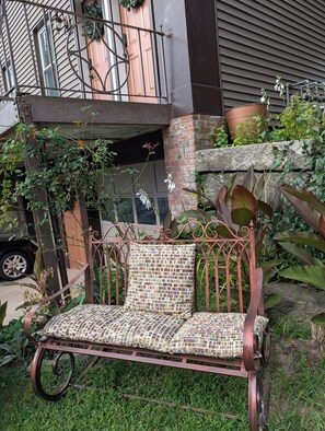 A wrought iron bench with a mosaic cushion in a garden area with potted plants and a building in the background.