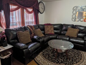A living room with a black leather sofa, a glass coffee table, a patterned rug, and a wall clock.