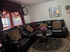 Una sala de estar con un sofá de cuero negro, almohadones estampados, una mesa de centro de vidrio y un espejo decorativo en la pared.