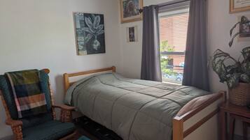 A single bed with a wooden frame, a chair with a plaid cushion, and a window with curtains.