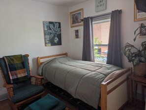 A single bed with a wooden frame, a chair with a plaid cushion, and a window with curtains.