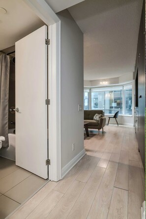 Un interior modern amb una porta corredissa que dona a un bany, una zona de seients amb un sofà i una cadira, i una cuina visible a través de portes de vidre.
