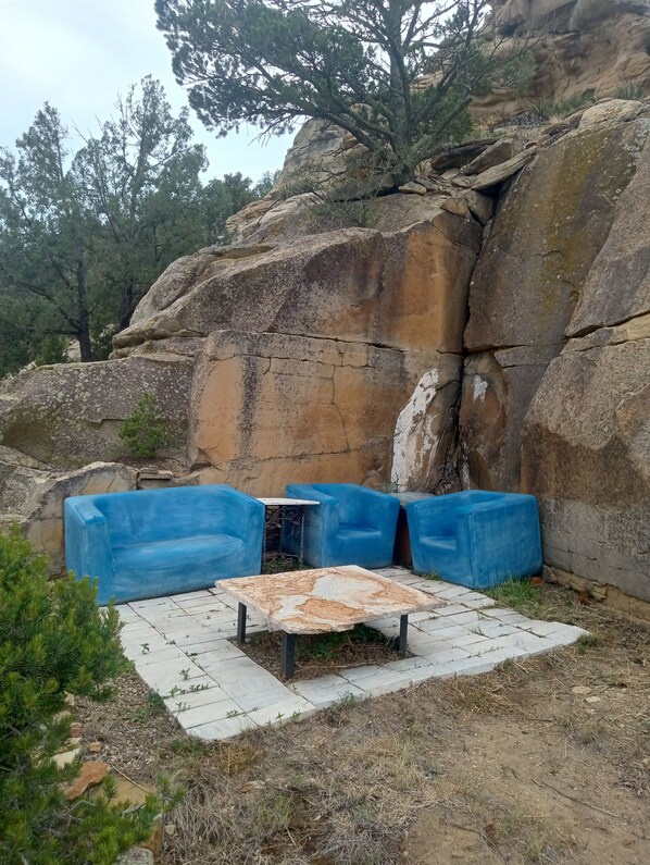 Un área de estar con bancos acolchados azules y una mesa de piedra, rodeada de formaciones rocosas naturales y vegetación escasa.