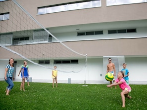 Eine Gruppe von Menschen spielt Volleyball auf einer Wiese, im Hintergrund ist ein Gebäude zu sehen.