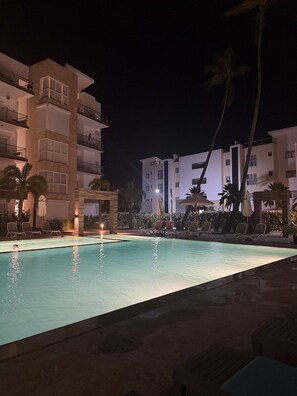 A swimming pool with lounge chairs and palm trees, surrounded by multi-story buildings with balconies. - Navío Pool-Front Patio · Beach Access · Power Incl (Punta Cana)