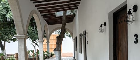 Un paseo techado con suelo de piedra, vigas de madera, plantas en macetas y un banco.