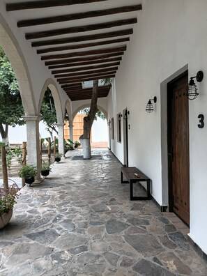 A covered walkway with stone flooring, wooden beams, potted plants, and a bench.