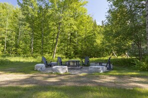 An outdoor seating area with Adirondack chairs and a fire pit surrounded by rocks and trees. - Bright Condo | Bike+ Randos In/Out • Vue Montagne (Les Laurentides Regional County Municipality)