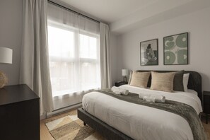 A bedroom with a large bed, a bedside table, a lamp, and a window with sheer curtains. - Bright Condo | Bike+ Randos In/Out • Vue Montagne (Les Laurentides Regional County Municipality)