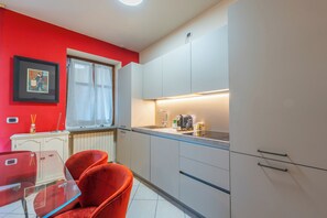 Una cocina moderna con armarios blancos, pared roja, ventana con cortinas y comedor con mesa de vidrio y sillas rojas.
