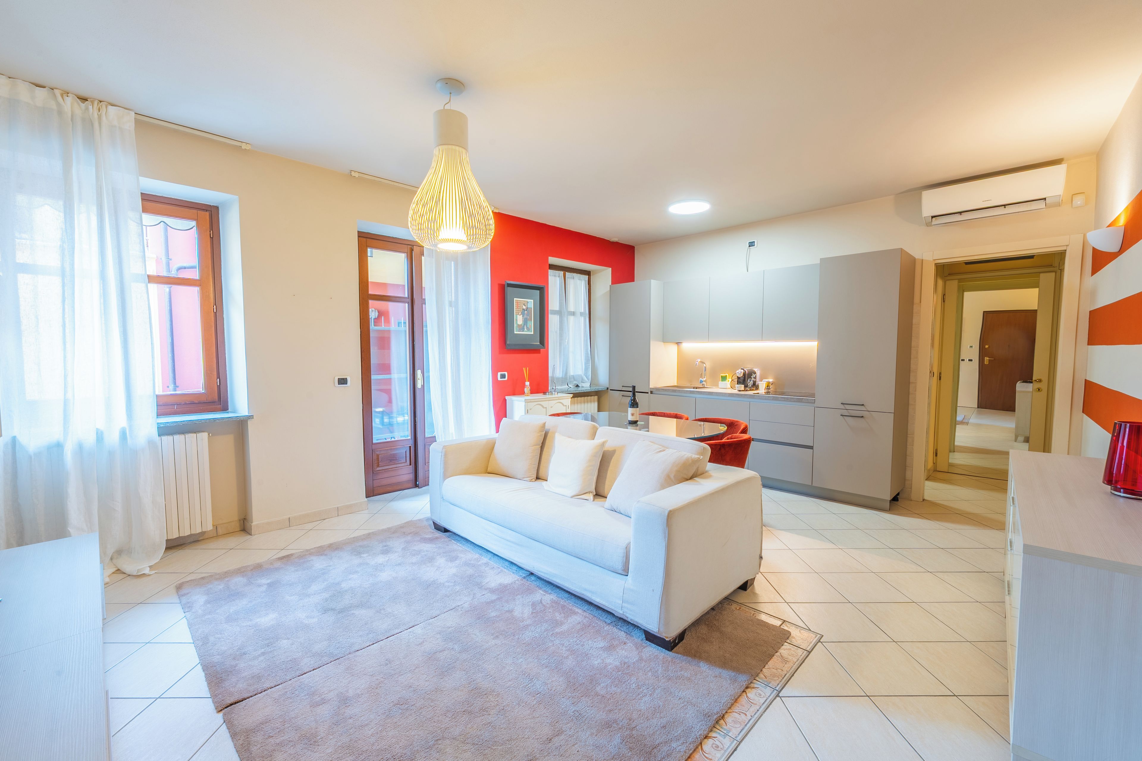 A modern living room with a white sofa, a kitchen area with white cabinets, and a dining table with red chairs.