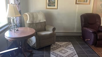 Een woonkamer met een ronde houten tafel, een fauteuil met kussens, een kamerkleed met patroon en twee ingelijste foto's aan de muur.