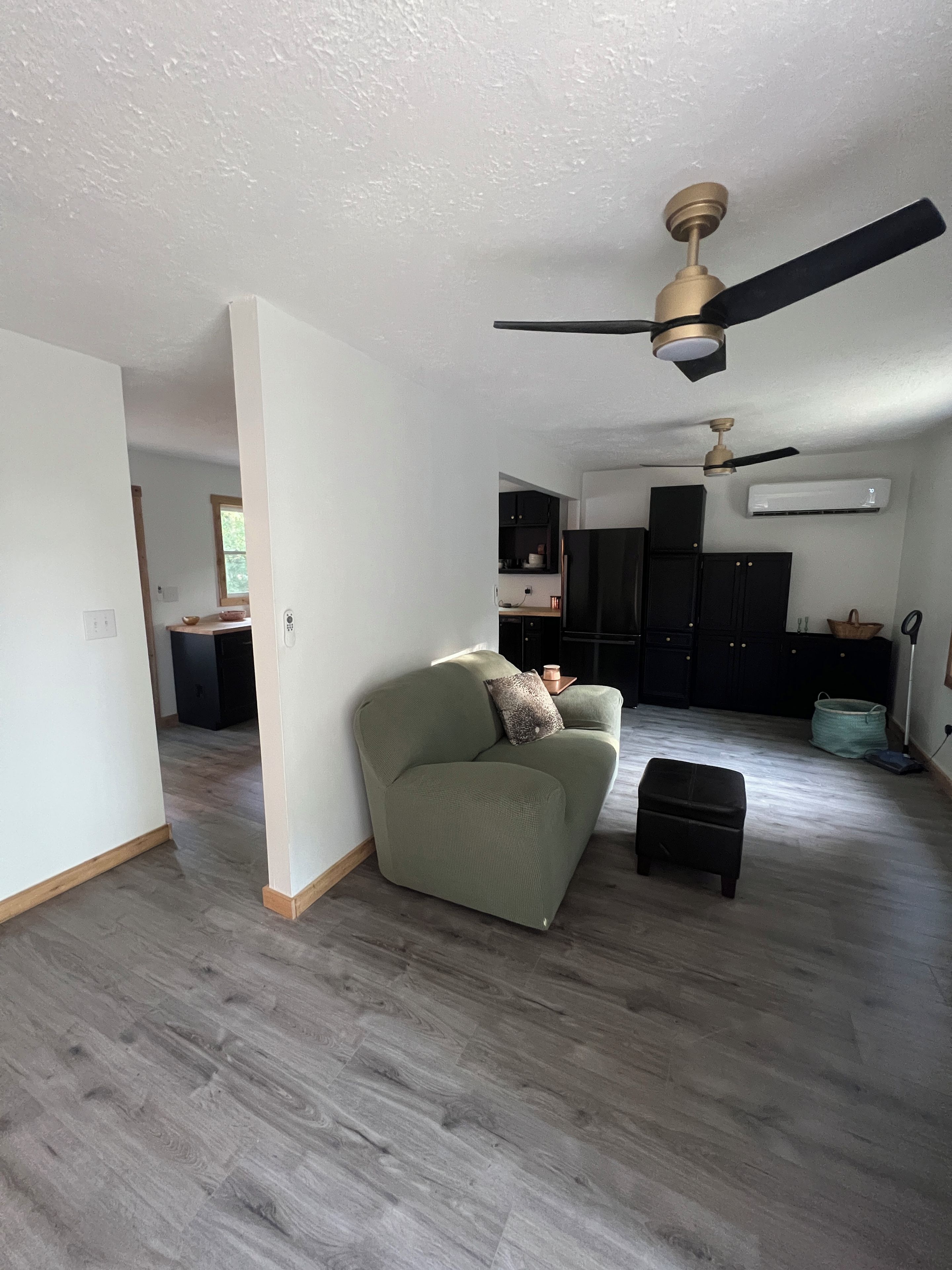 A spacious living area with a sofa, ottoman, and ceiling fan. A kitchen with cabinets and an air conditioning unit is visible in the background.