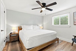 Ein Schlafzimmer mit Bett, Nachttisch, Lampe, Deckenventilator, Fenster mit Jalousien und Holzboden.