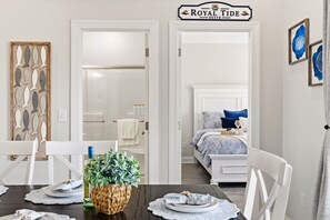 A hotel room with a bed, a dining table with chairs, a bathroom with a shower, and a sign above the door reading 'The Royal Tide Suite'.