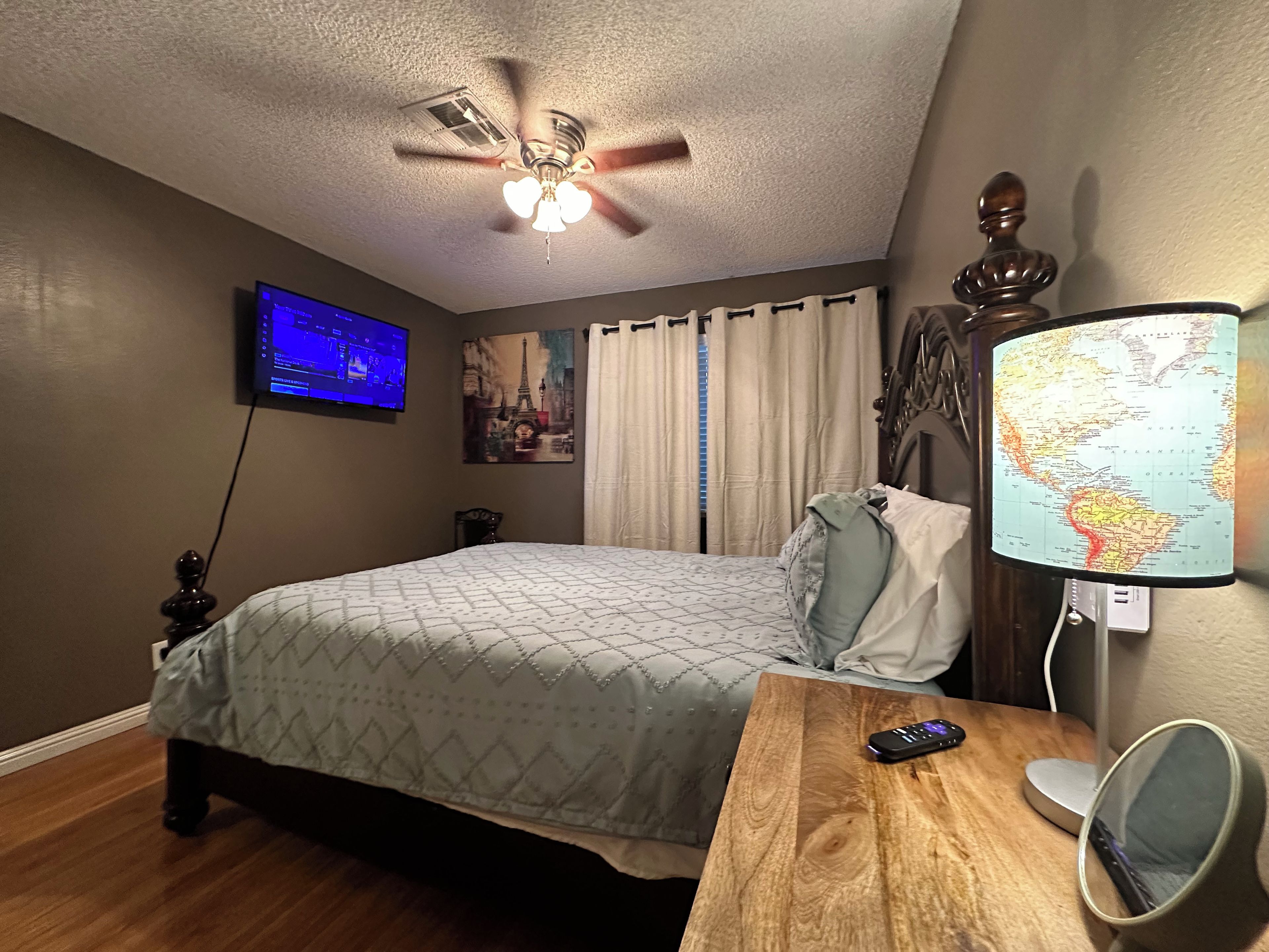 A bedroom with a bed, a ceiling fan, a TV, a map lamp, and a wooden nightstand.