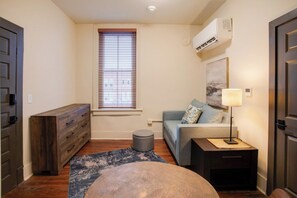 A compact living space with a sofa, a small ottoman, a side table with a lamp, a dresser, and a window with blinds. - Downtown Florence Retreat | Walkable Historic Stay (Florence)