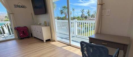 A room with a wooden floor, a TV mounted on the wall, a desk, a chair, and a view of palm trees through a sliding glass door.
