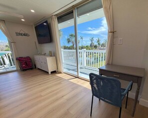 Ein Zimmer mit Holzboden, einem an der Wand montierten Fernseher, einem Schreibtisch, einem Stuhl und Blick auf Palmen durch eine Schiebetür.
