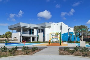 Ein modernes Gebäude mit einem Schwimmbad, einem Spielplatz mit bunten Geräten und blauem Himmel.