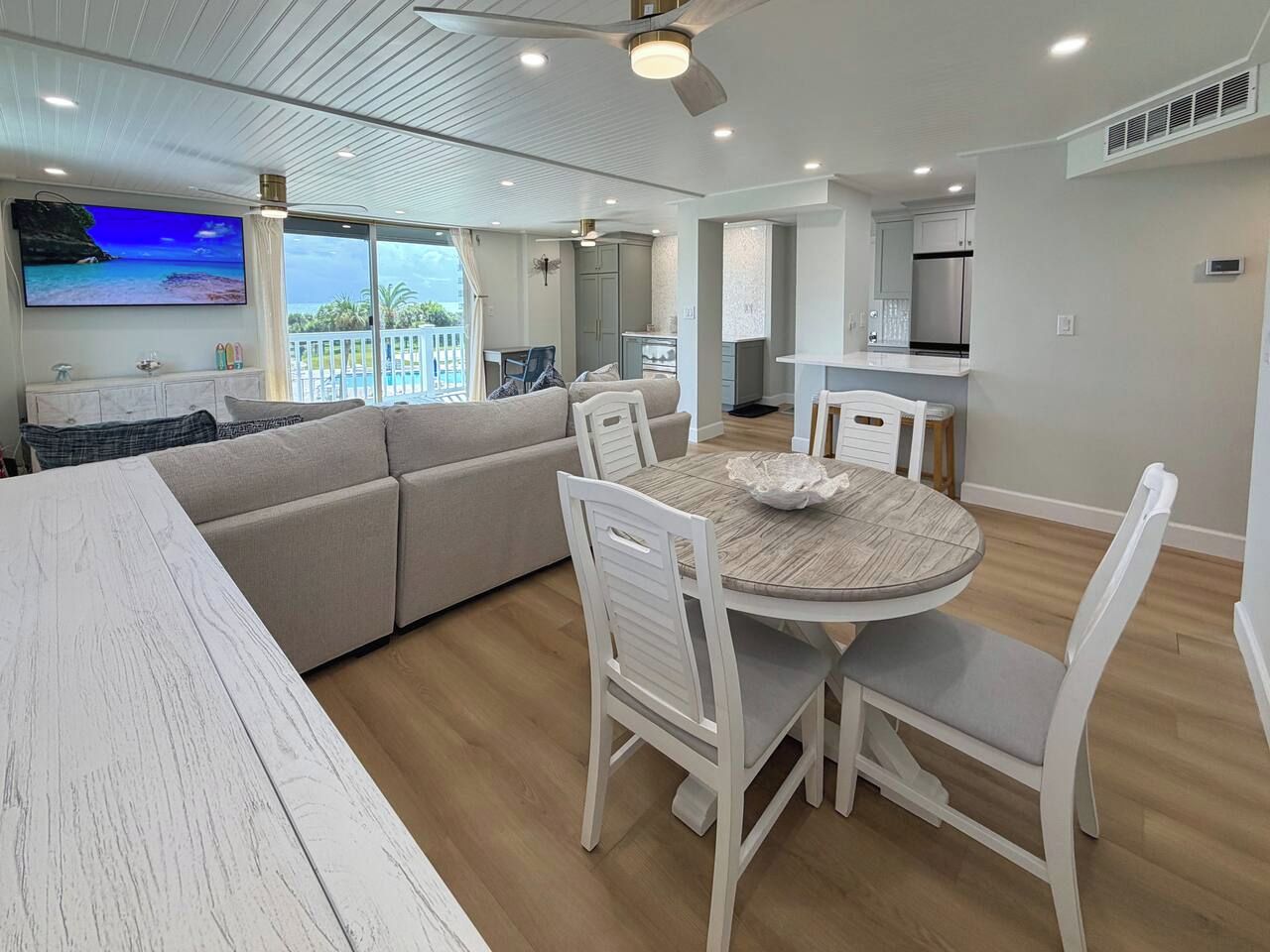 A modern living room with a round dining table, white chairs, a sofa, and a TV displaying a beach scene.