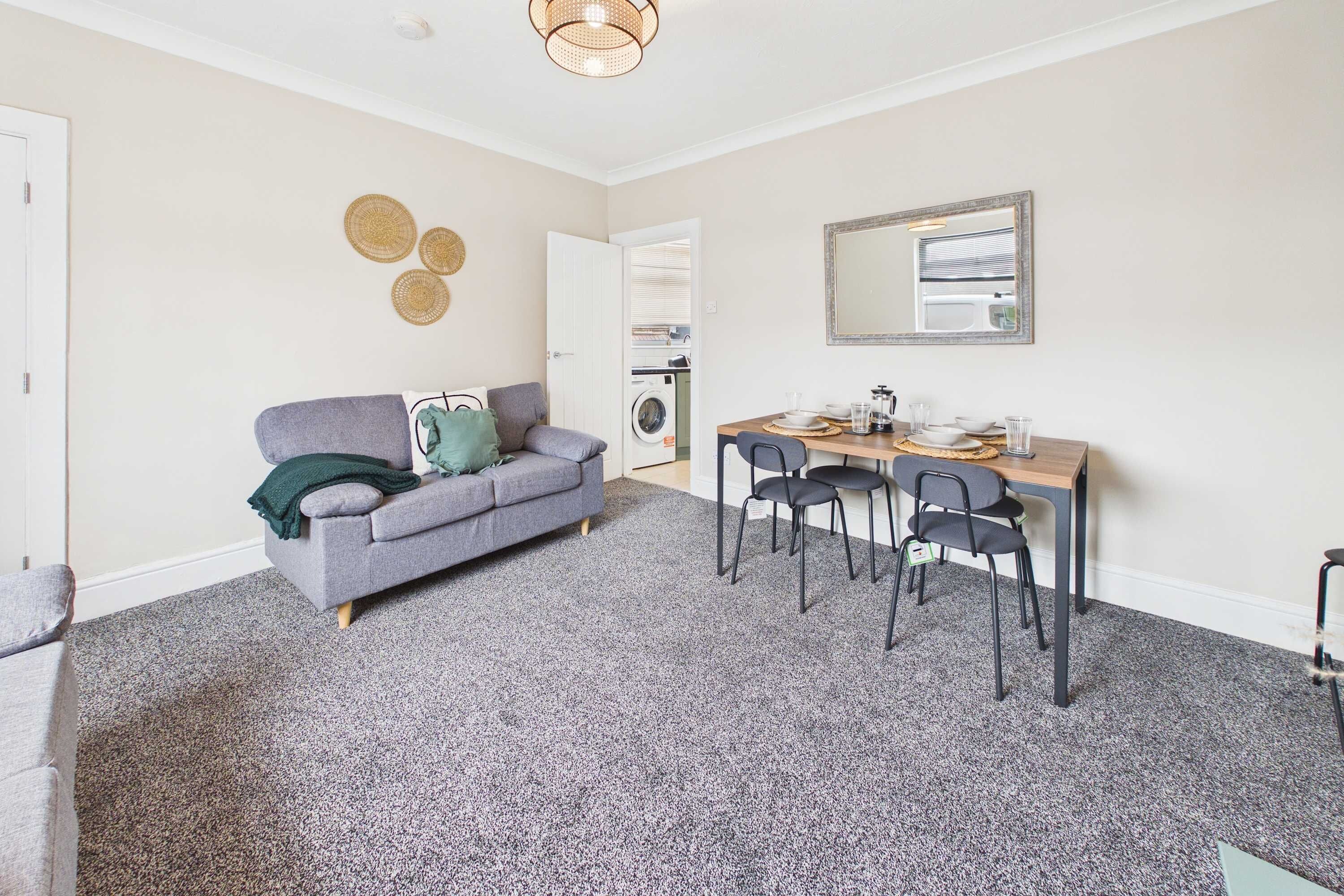 A compact living space with a grey sofa, a dining table with chairs, and a washing machine in the background.