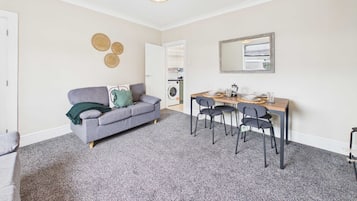 A compact living space with a grey sofa, a dining table with chairs, and a washing machine in the background.