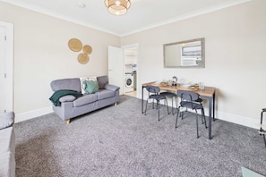 Un espace de vie compact avec un canapé gris, une table à manger avec des chaises, et un lave-linge à l’arrière-plan.