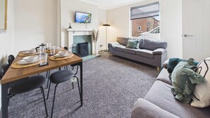 Een woonkamer met een open haard, een eetgedeelte met een tafel voor vier personen, en een bank met kussens.